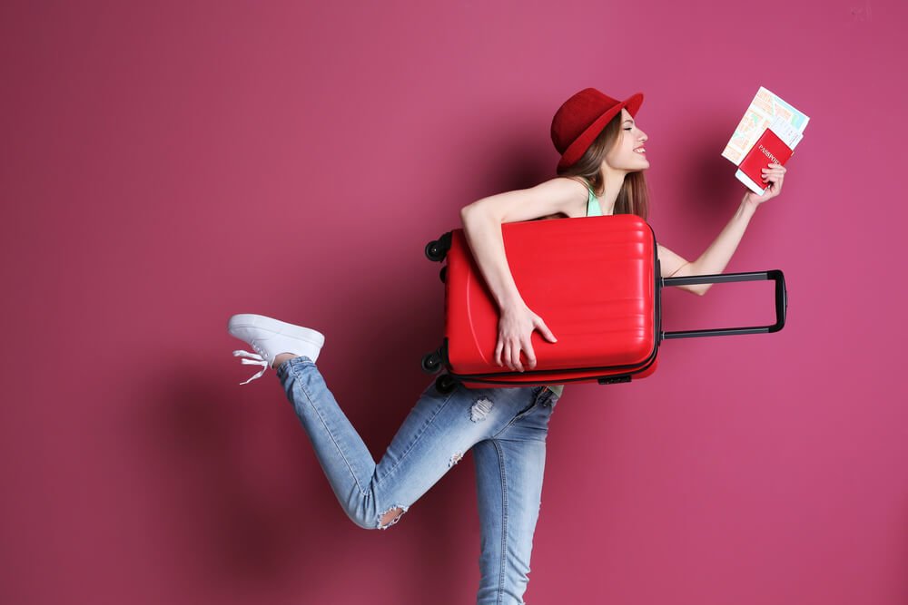 ピンクの背景の前で赤いスーツケースを小脇に抱える女性.jpg